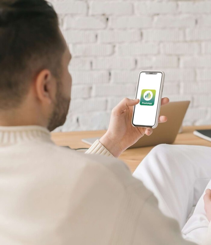 Ein Mann sitzt am Tisch und blickt auf das Smartphone in seiner Hand – er öffnet gerade die Praxis-App.