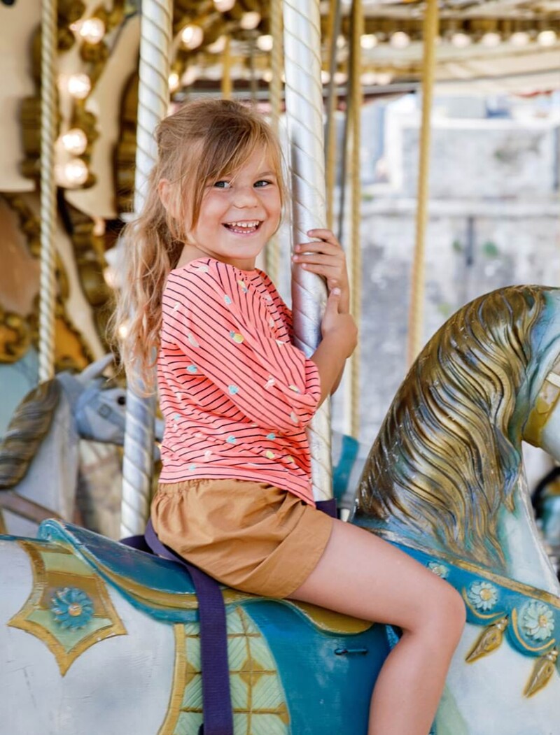 Mädchen sitzt in einem Retro-Karusell auf einem Pferd und schaut glücklich grinsend in die Kamera, während sie darauf fährt.