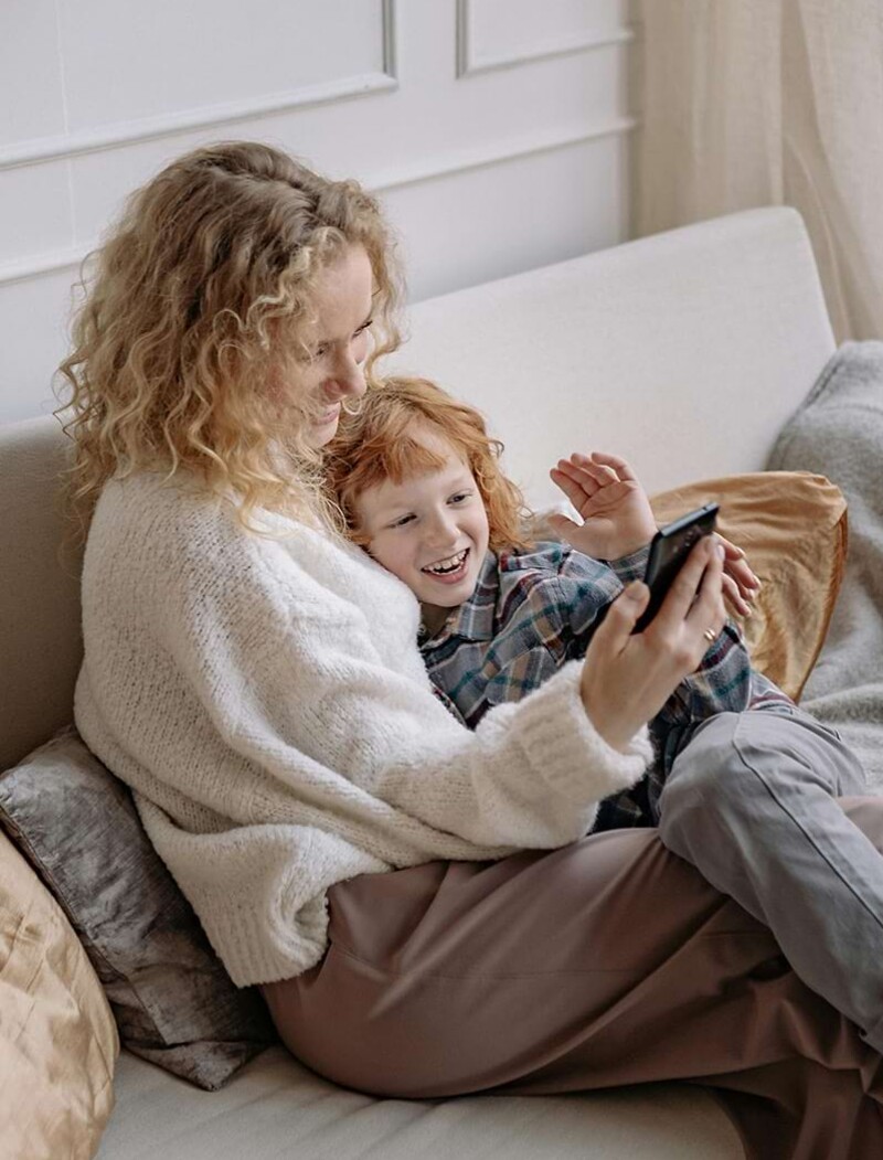 Eine Mutter sitzt kuschelnd mit ihrer Tochter auf dem Sofa. Sie beide schauen gemeinsam auf das Smartphone.