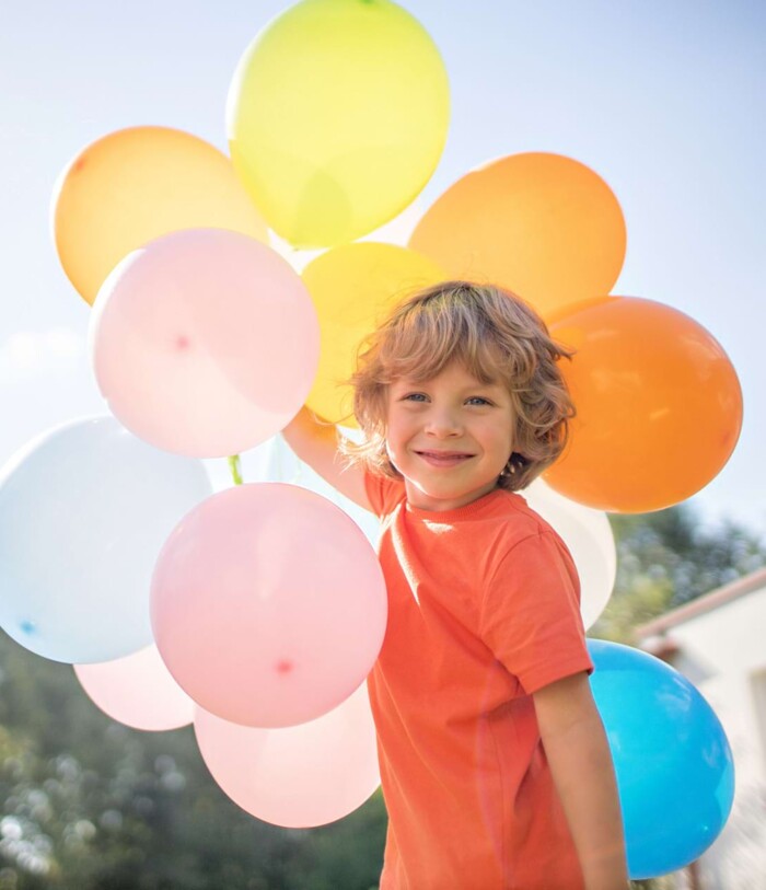 Ein blonder, lockiger Junge hält einen Strauß voll bunter Luftballons in der Hand und läuft durch den Garten.
